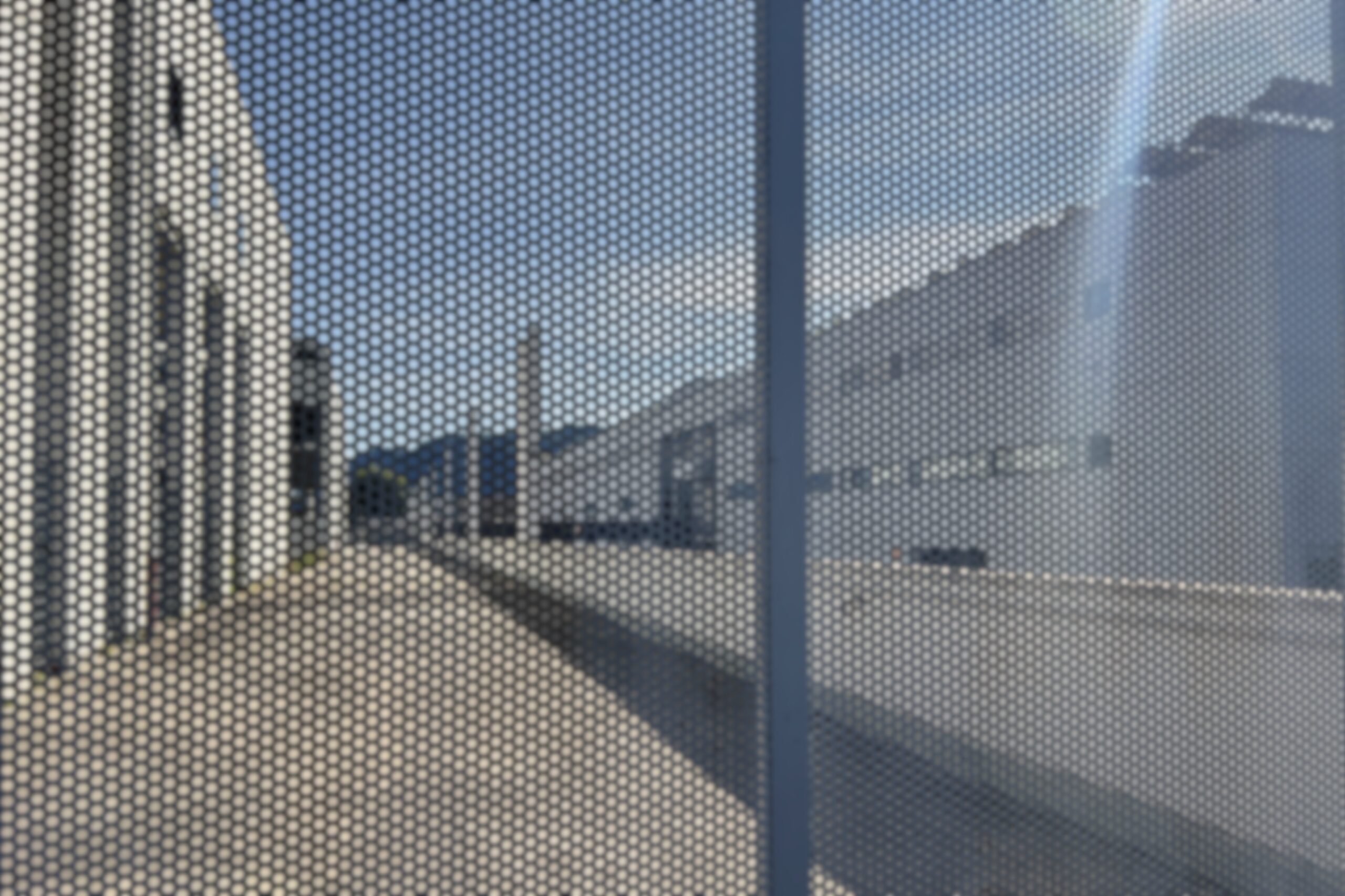 Perforated metal screen with a vertical steel bar in the foreground, revealing an industrial courtyard and buildings in soft, diffused light.